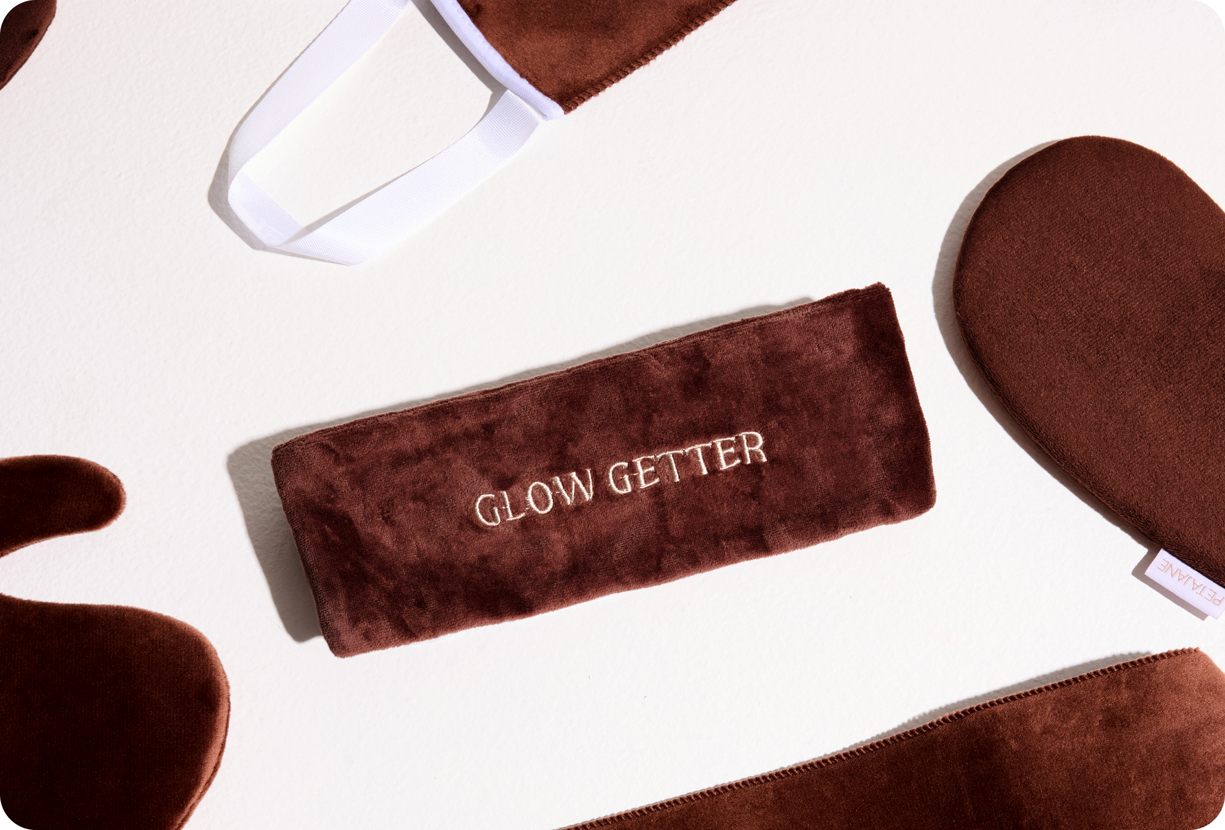 A flat lay of dark brown velvet tanning accessories on a white surface, featuring a headband embroidered with "GLOW GETTER" alongside a Peta Jane tanning mitt.