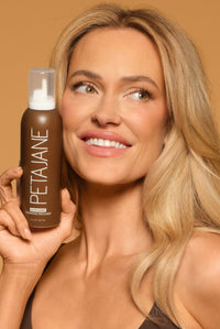 Founder Peta Murgatroyd smiling and holding a bottle of Peta Jane Beauty Ultra Dark Tanning Mousse against a warm tan background, showcasing a flawless sun-kissed glow.